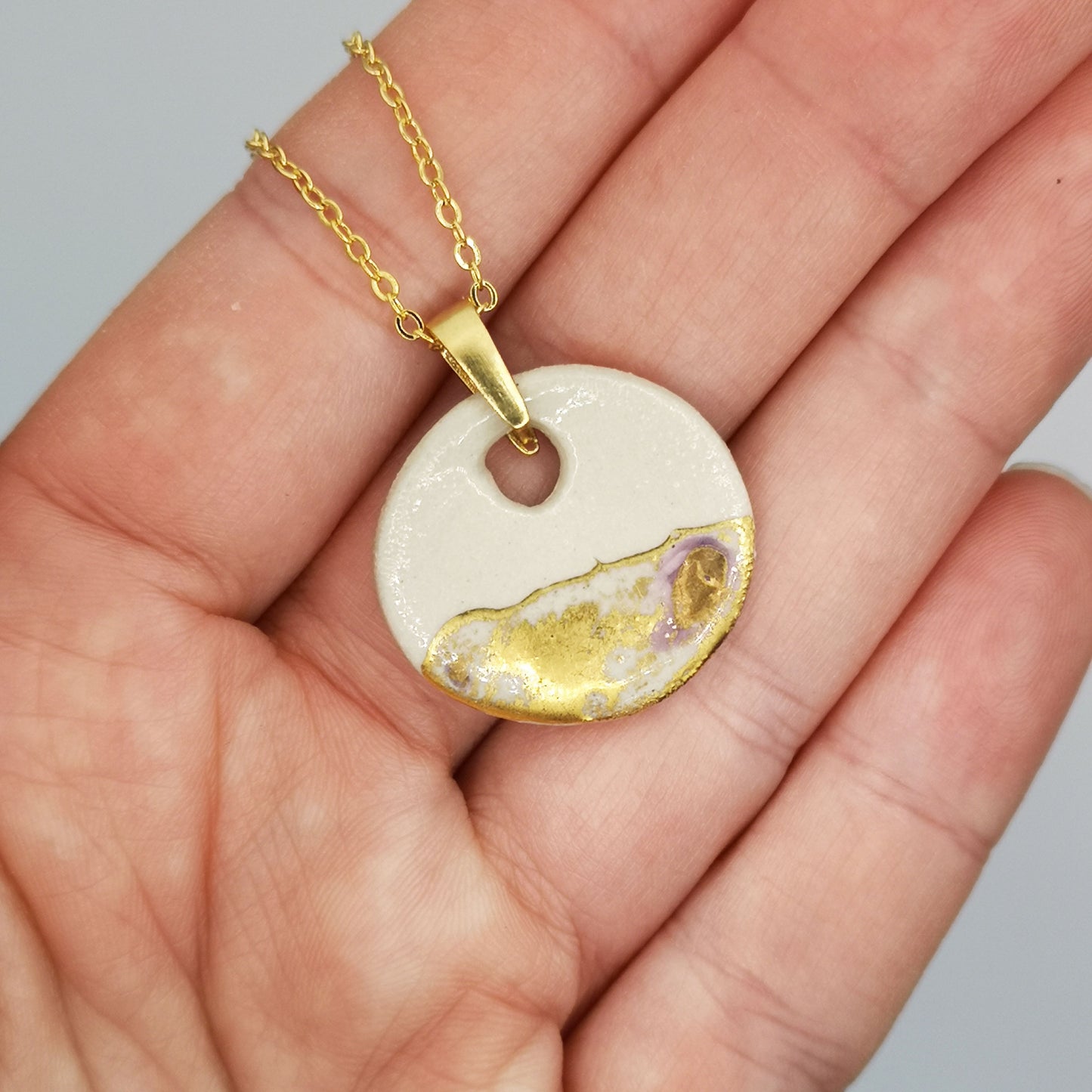 Gros plan sur un pendentif médaillon blanc en céramique avec détails dorés, suspendu à une chaîne en or fin.