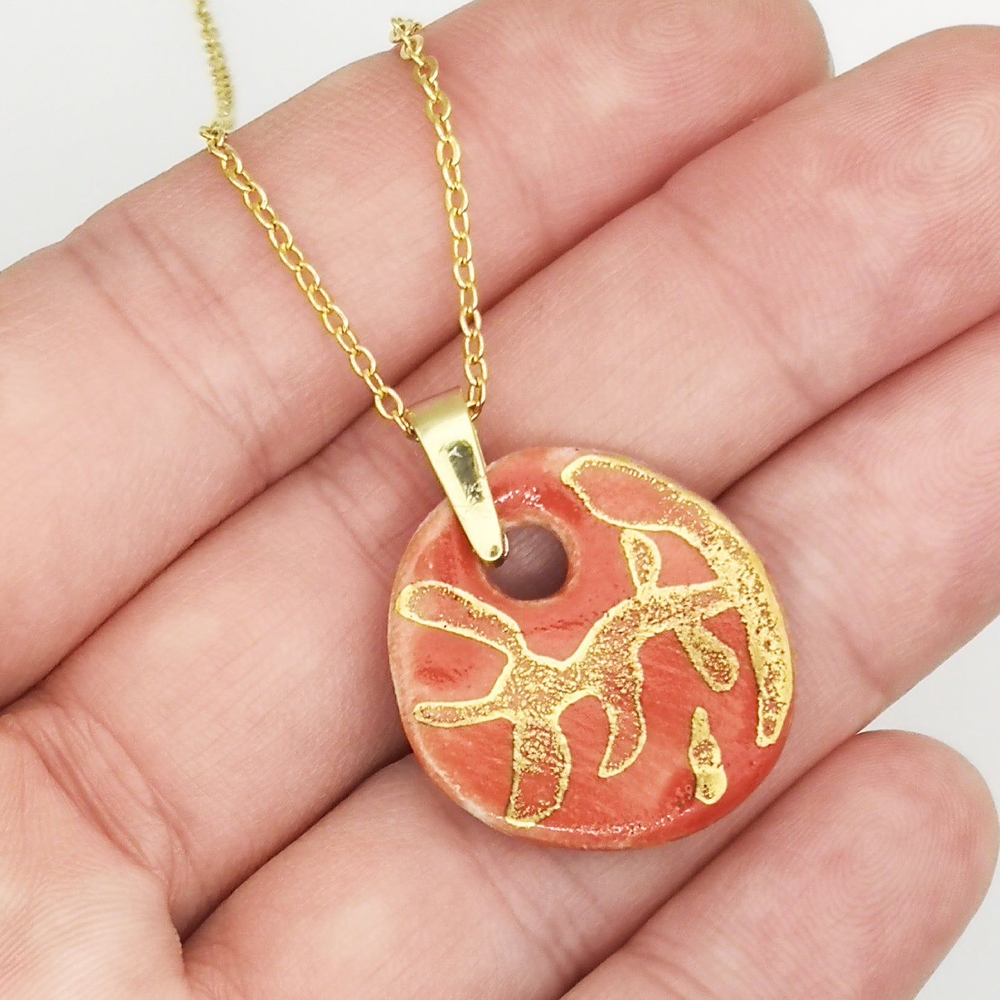 Collier en céramique rouge corail avec un médaillon artisanal et des détails dorés inspirés de la nature, dans un écrin rose prêt à offrir.