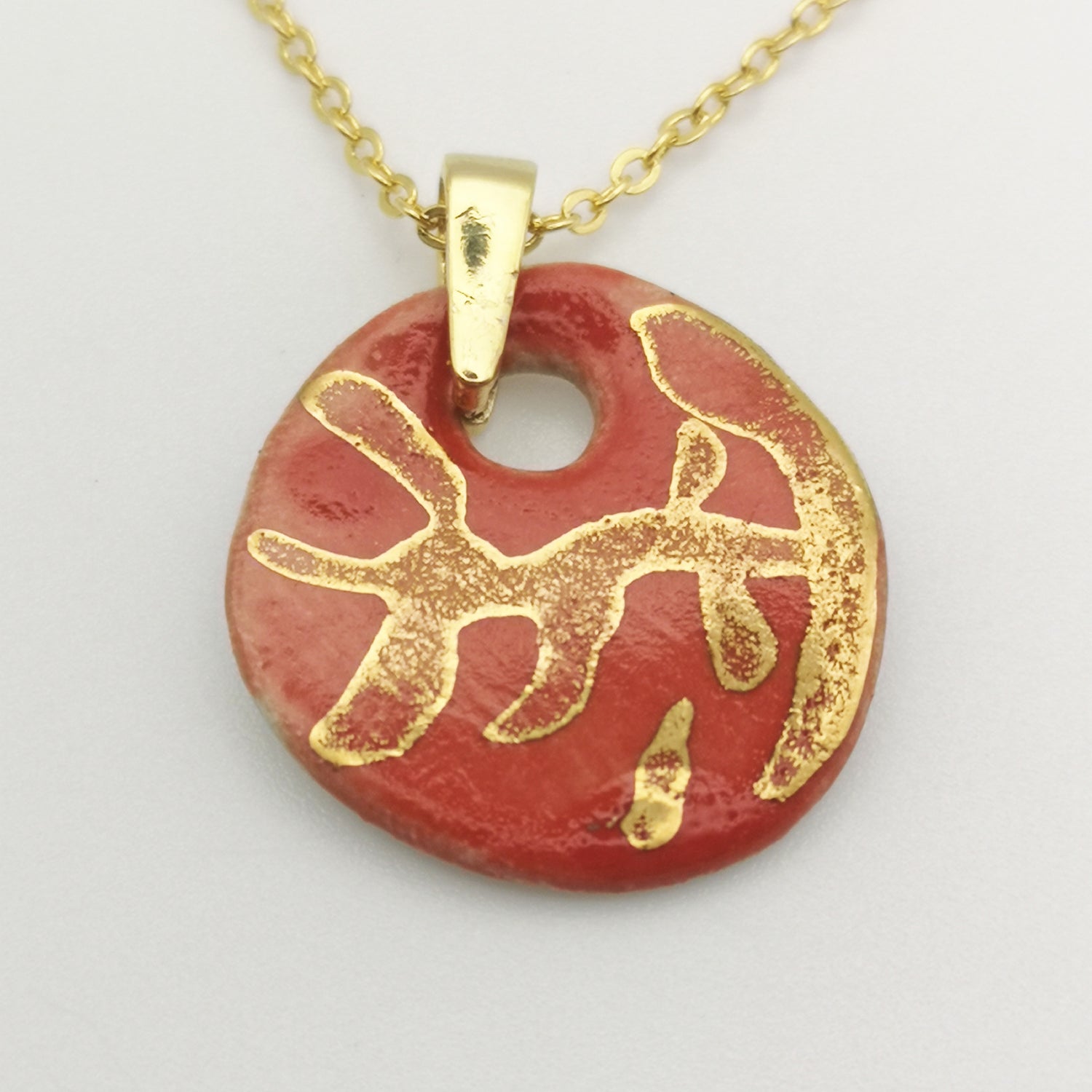 Gros plan sur un pendentif en céramique rouge corail avec des branches dorées peintes à la main, suspendu à une chaîne dorée en laiton.